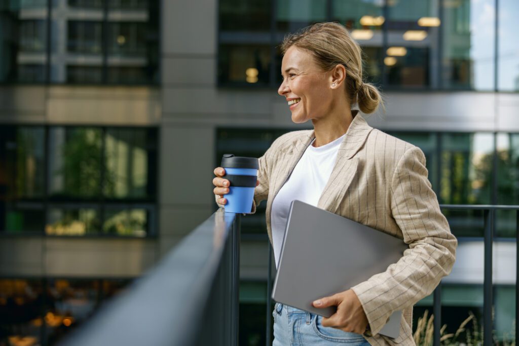 Sales manager standing with laptop and coffee on office terrace during break time and looks away